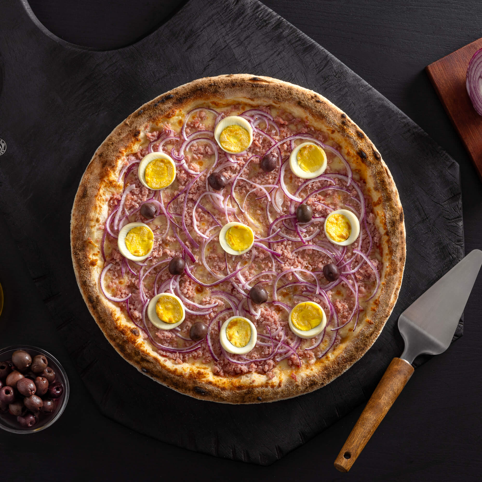 Pizza portuguesa com massa de fermentação lenta de 48 horas, molho artesanal, queijo derretido, presunto, cebola, ovos e azeitonas.